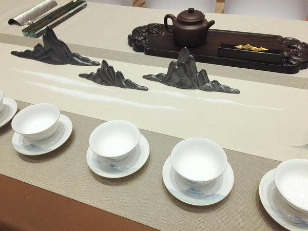 A tea table at the First Exchange of Tea Culture Between Beijing and Taiwan, Beijing, June 24, 2018. (Photo provided to chinadaily.com.cn)
An exquisite porcelain teapot. Several fine ceramic cups. A small vase and a flower. A piece of calligraphy introducing tea making procedures.
These fives things adorn a traditional Chinese tea table. With a touch of Chinese aesthetic, the table is almost an artwork in the form of modern installation.
Another piece was inspired by A Thousand Miles of Rivers and Mountains, a masterpiece by Song Dynasty (960-1279) painter Wang Ximeng. The painting was featured on cups and table flag which were decorated with black mountain-shaped sculptures.
A total of 30 such tables, from both Chinese mainland and Taiwan, were featured at the First Exchange of Tea Culture Between Beijing and Taiwan at Grand Millennium Beijing on June 24.